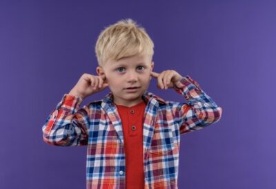 a little boy with blonde hair wearing checked shirt keeping hand on ears on a purple background. Sensory Integration Challenges in Children: Insights and Strategies from Red Door Pediatric Therapy